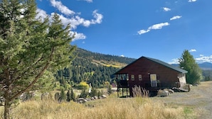 A wooden cabin with a porch, surrounded by a grassy field and trees, with a mountain in the background.