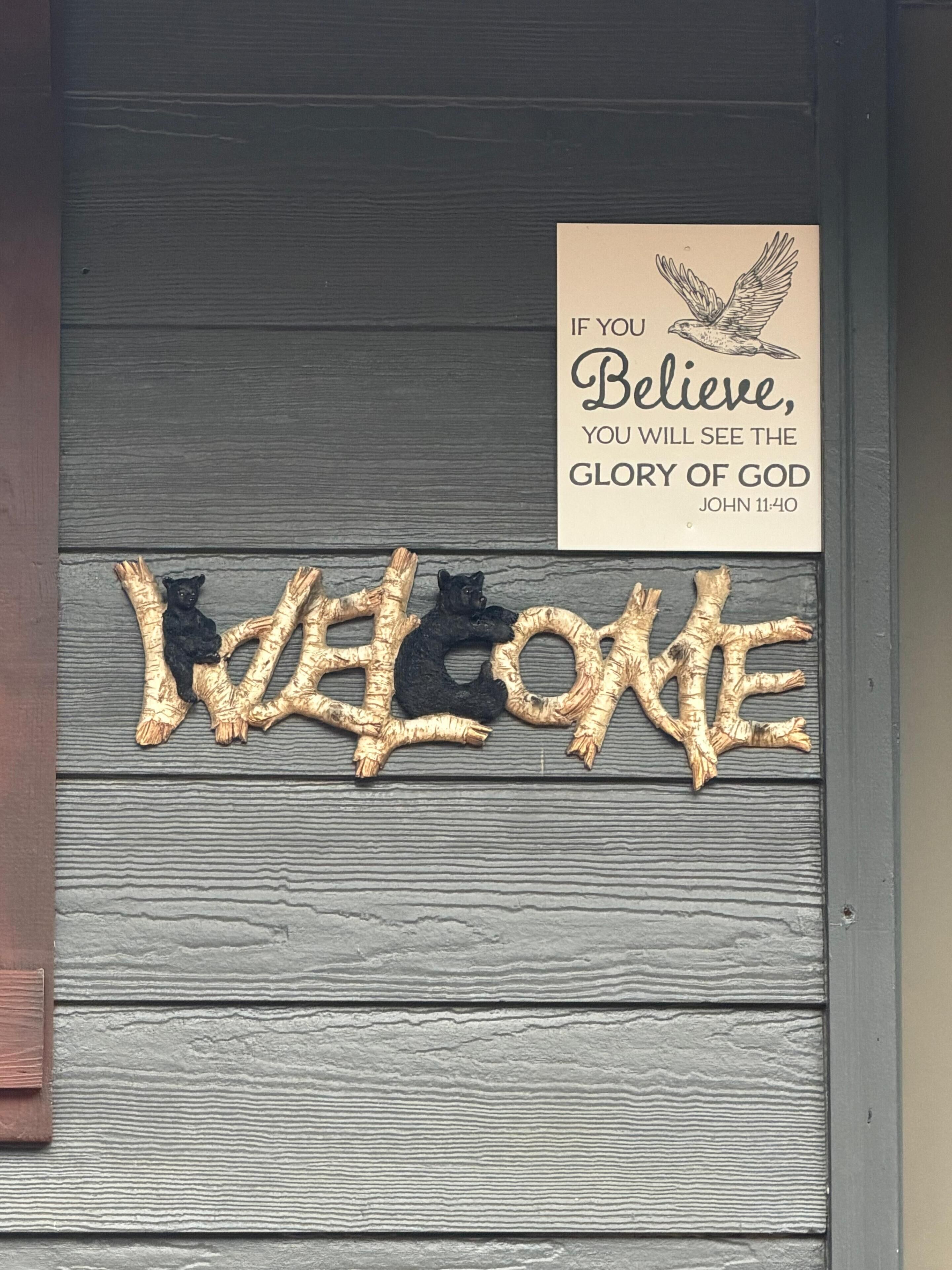 Een houten deur met een bord waarop staat 'IF YOU BELIEVE, YOU WILL SEE THE GLORY OF GOD' en een decoratief 'WELCOME'-bord.