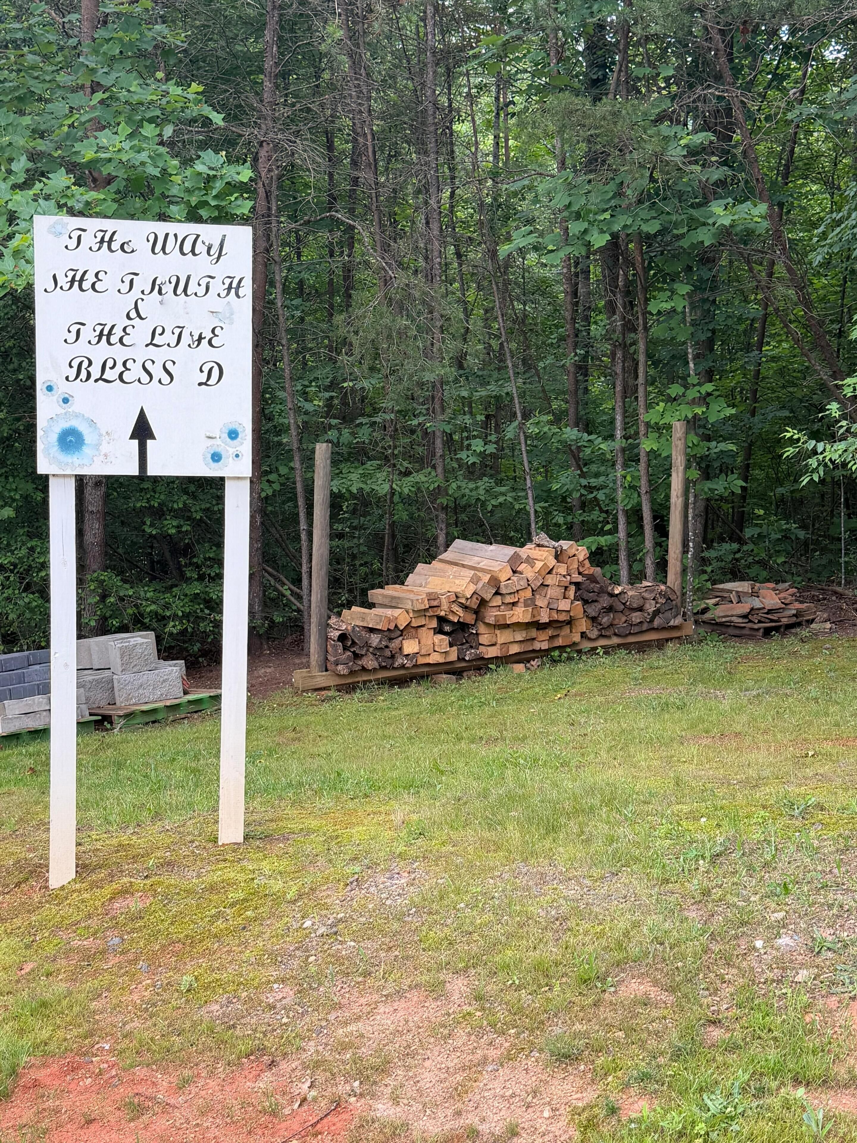 A sign with text and an arrow, surrounded by a forest and stacks of wood.