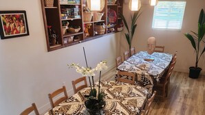 Ein Essbereich mit einem langen Tisch, bedeckt mit einer gemusterten Tischdecke, Holzsesseln, hängenden Pendelleuchten, einem Blumentopf und einem Regal mit diversen Gegenständen.