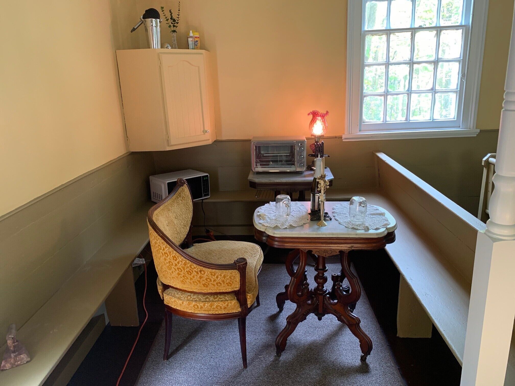 A corner room with a vintage chair, a table with candles, a microwave, and a window with a view.