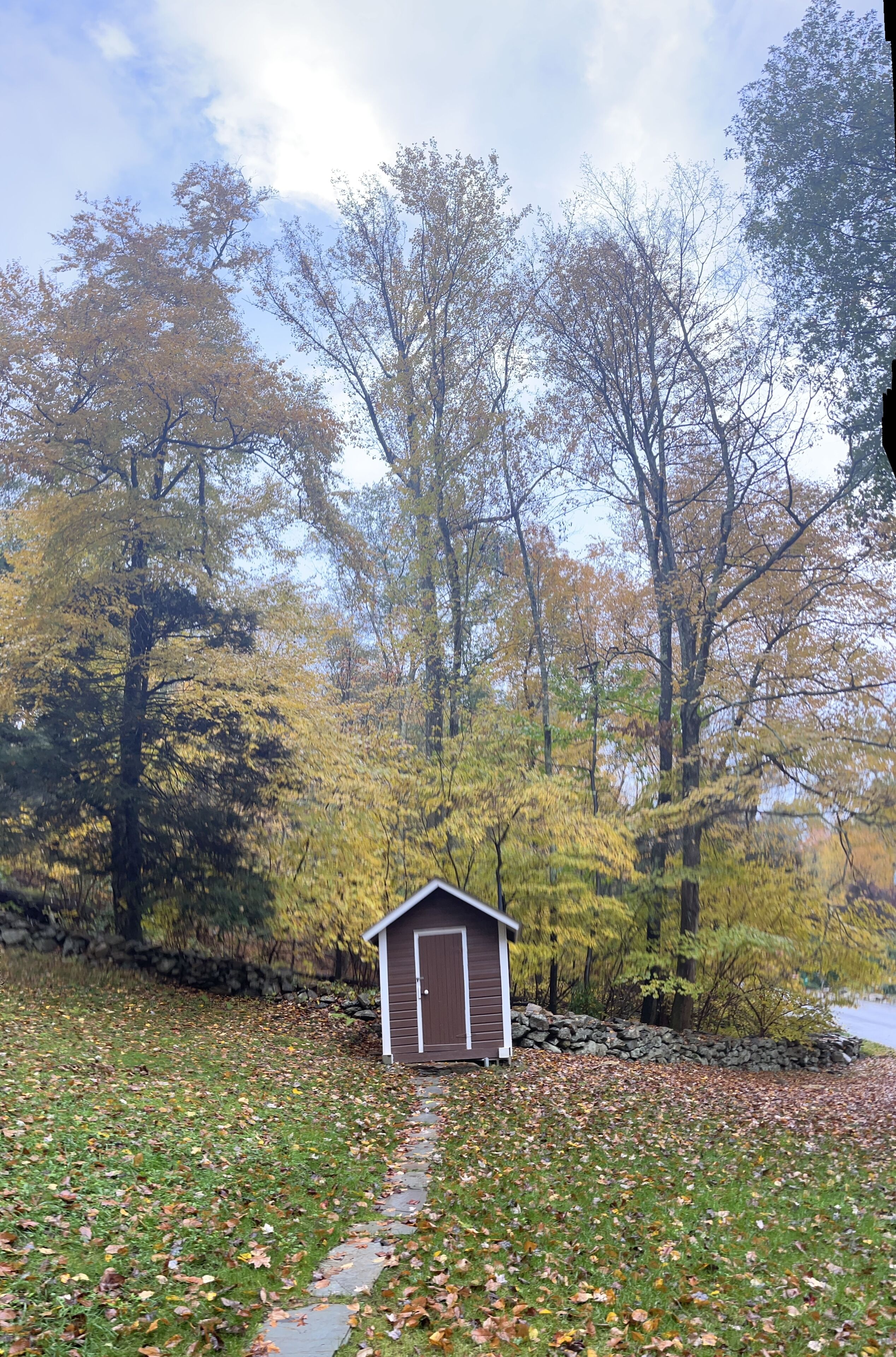 A small wooden shed in a wooded area with a stone wall and a path leading to it.