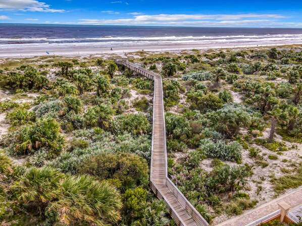 A wooden boardwalk extending through a coastal area with dense vegetation and a beach in the background.