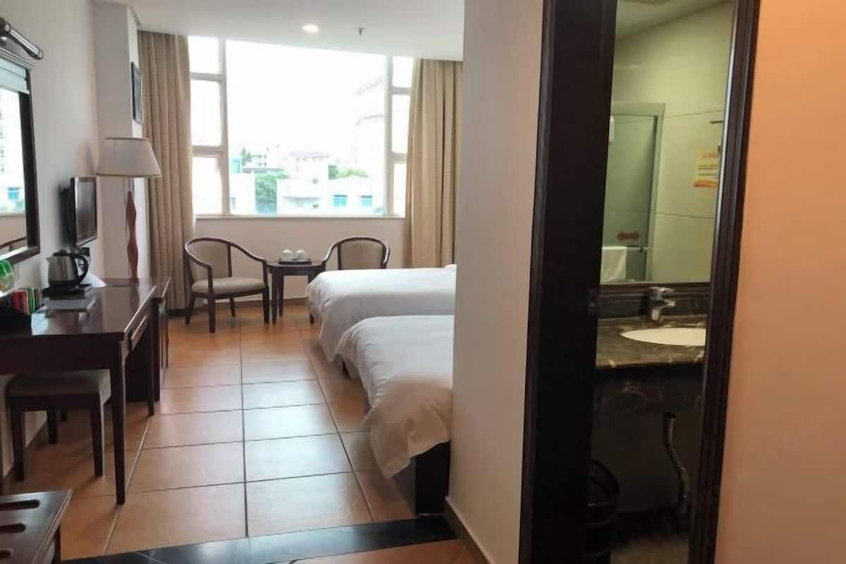 A hotel room with a bed, a desk with a lamp, a chair, a small table, and a bathroom with a sink and mirror.