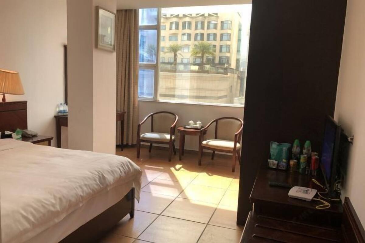 Ett hotellrum med en säng, två stolar, ett litet bord, en tv och ett fönster med utsikt över byggnader.