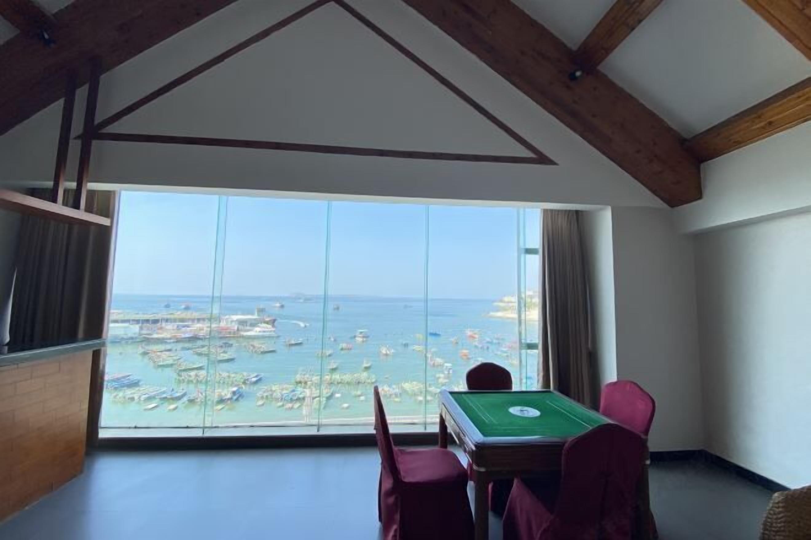 A room with a pool table, large windows with a view of boats and a beach, and wooden beams on the ceiling.