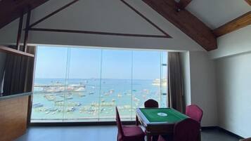 A room with a pool table, large windows with a view of boats and a beach, and wooden beams on the ceiling.
