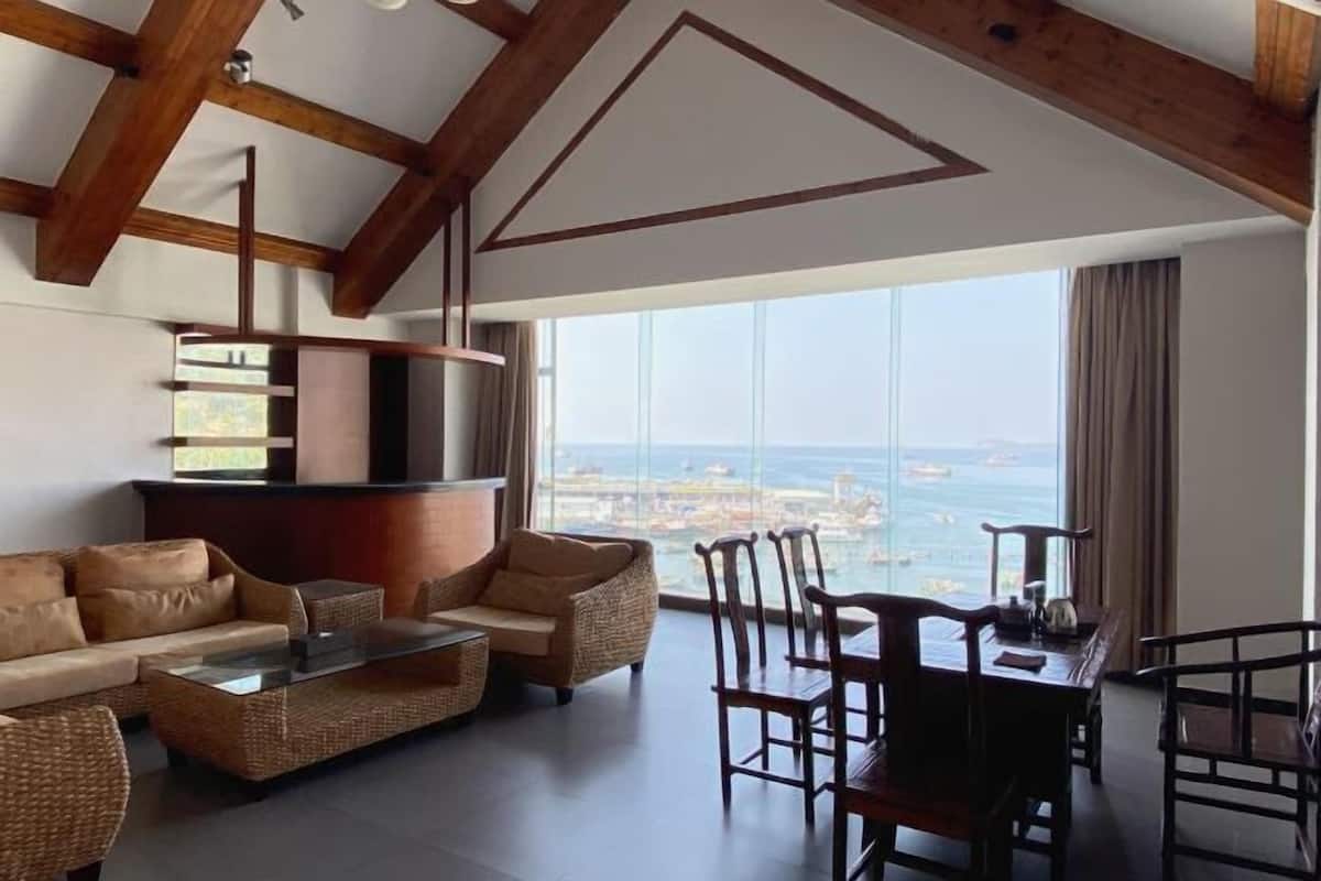A spacious living room with a large window offering a view of the sea and cityscape. There is a sofa, a coffee table, and wooden chairs.