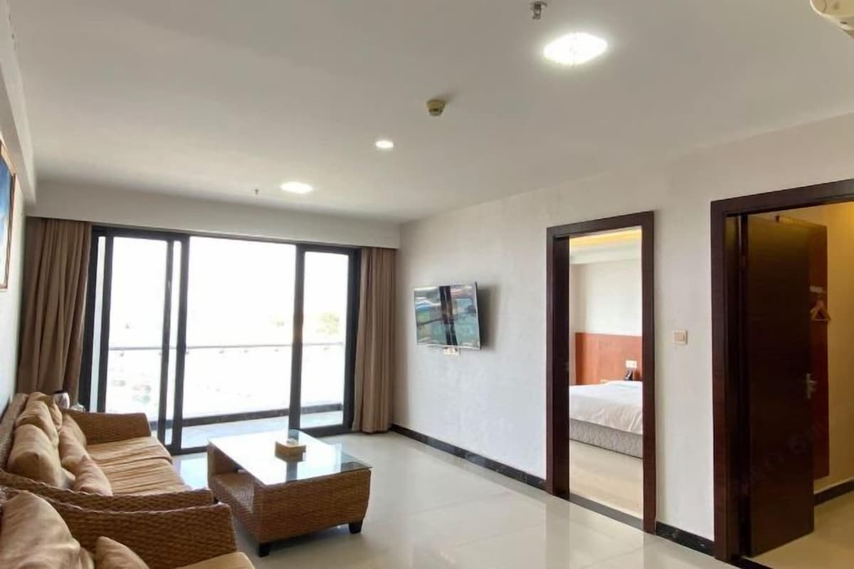 A hotel room with a sofa set, a coffee table, a TV mounted on the wall, and a bed with a headboard.