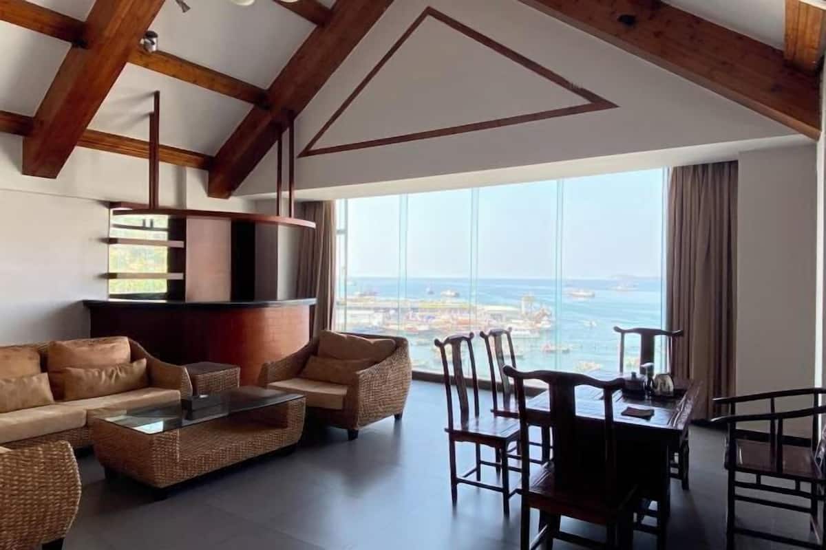 A spacious living area with a high ceiling, wooden beams, a large window with a view of the sea, a sofa, a coffee table, and a dining table with chairs.