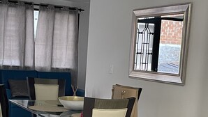 Un coin repas comprenant une table rectangulaire, quatre chaises, un canapé bleu et une fenêtre ornée d’une grille métallique.