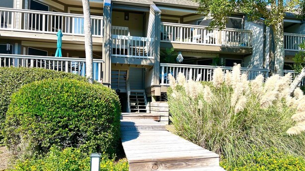 A multi-story building with a wooden deck leading to the entrance, surrounded by greenery and a clear sky. - Courtside Paradise by Sandpiper | Near Beach (Seabrook Island)