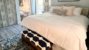 A bedroom with a large bed, a wooden bench, a chair, and a view of the outdoors. - Courtside Paradise by Sandpiper | Near Beach (Seabrook Island)