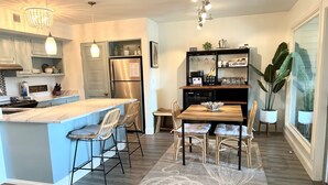 A modern kitchen with a central island, bar stools, a dining table with chairs, a coffee station, and a large potted plant. - Courtside Paradise by Sandpiper | Near Beach (Seabrook Island)