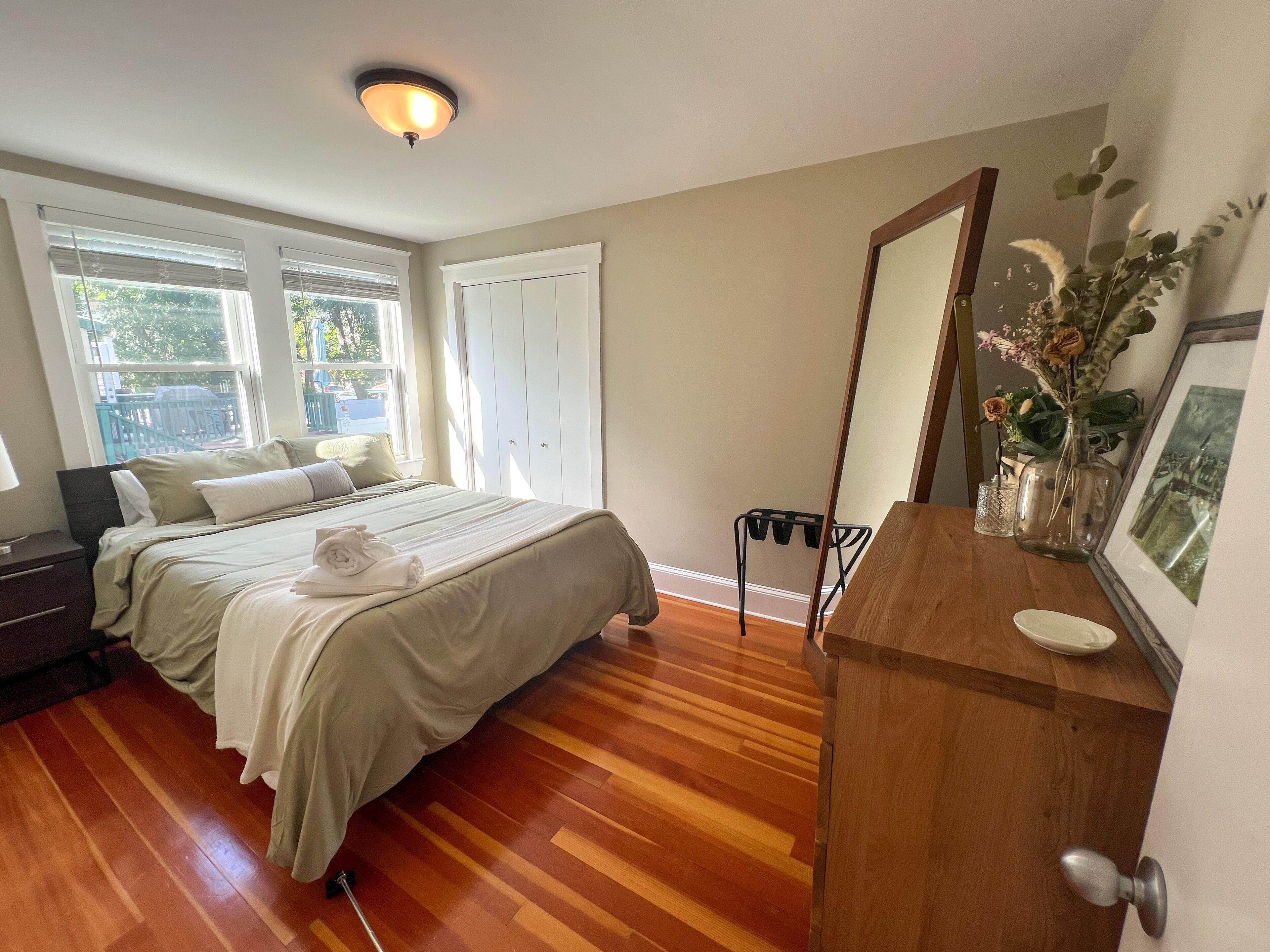 A bedroom with a bed, wooden floor, a dresser, a mirror, and a window with blinds.