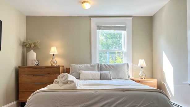 Un dormitorio ordenado con cama, mesitas de noche, cómoda y ventana que proporciona luz natural.