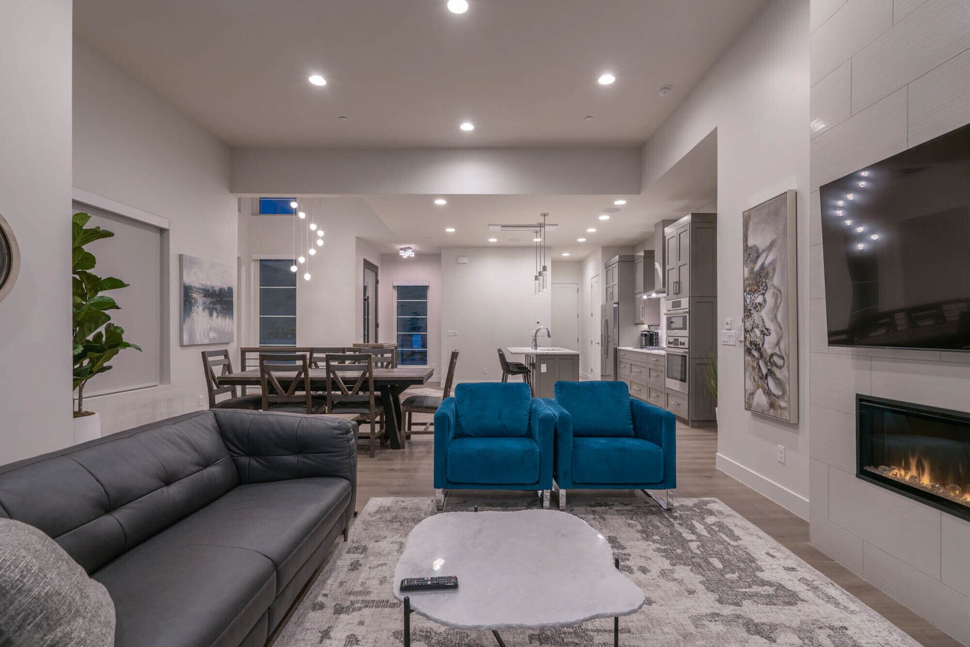 A modern living room with a gray sofa, a white coffee table, and blue armchairs. There is a fireplace on the right and a kitchen in the background.