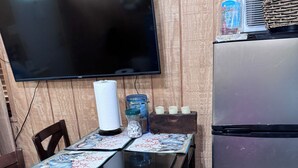 Una mesa de comedor de madera con cubierta de vidrio, un televisor montado en la pared y un refrigerador.