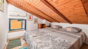 Un dormitorio con techo de madera inclinado, una cama con ropa de cama estampada, una ventana con marco azul y una mesita con una lámpara.