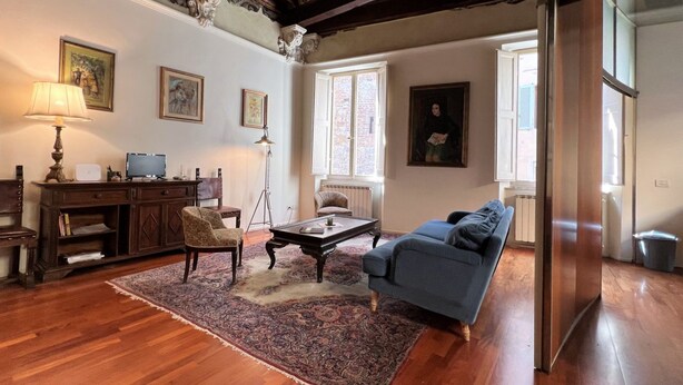 Een woonkamer met een blauwe bank, een houten salontafel en een tapijt met een patroon. Er staat een donker houten kast met een lamp, en er hangt een schilderij aan de muur.
