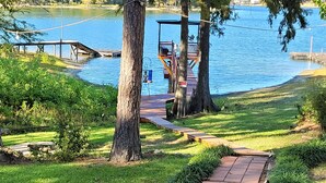 Una terraza de madera que conduce a un muelle sobre el agua, rodeada de césped y árboles.