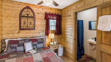 A wooden cabin room with a bed, a ceiling fan, a window with curtains, a small bedside table with a lamp, and a bathroom visible through an open door.