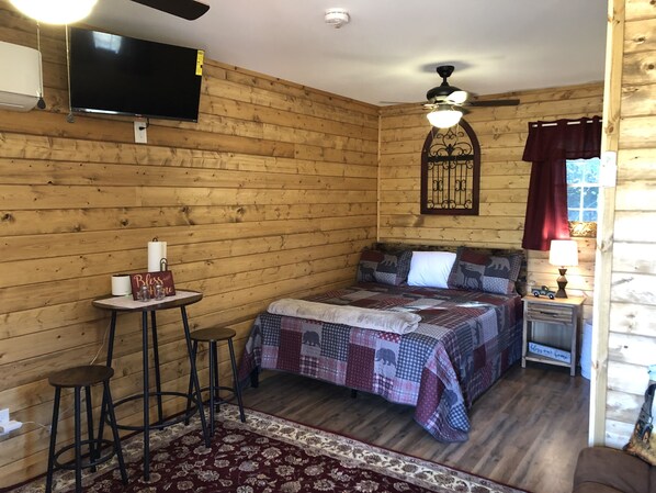 A cozy room with a bed, a small table with stools, a TV mounted on the wall, and a ceiling fan.