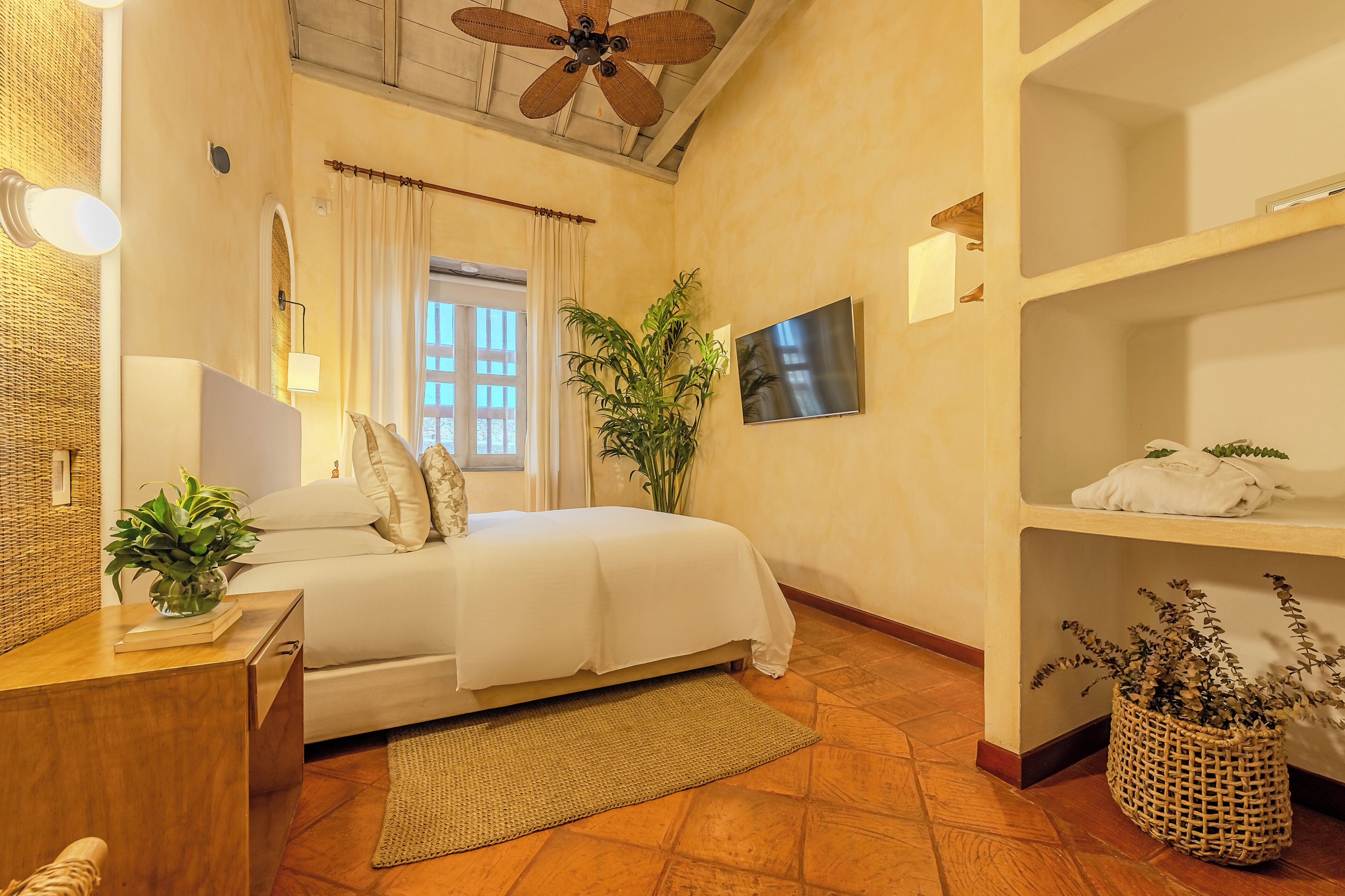 A bedroom with a bed, bedside table, lamp, TV, and a shelf with towels.