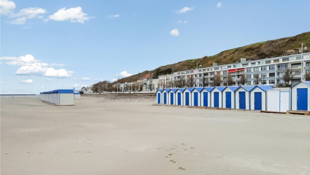 Une plage bordée d’une rangée de cabanes de plage bleues et blanches, avec, en arrière-plan, une rangée d’immeubles à plusieurs étages.