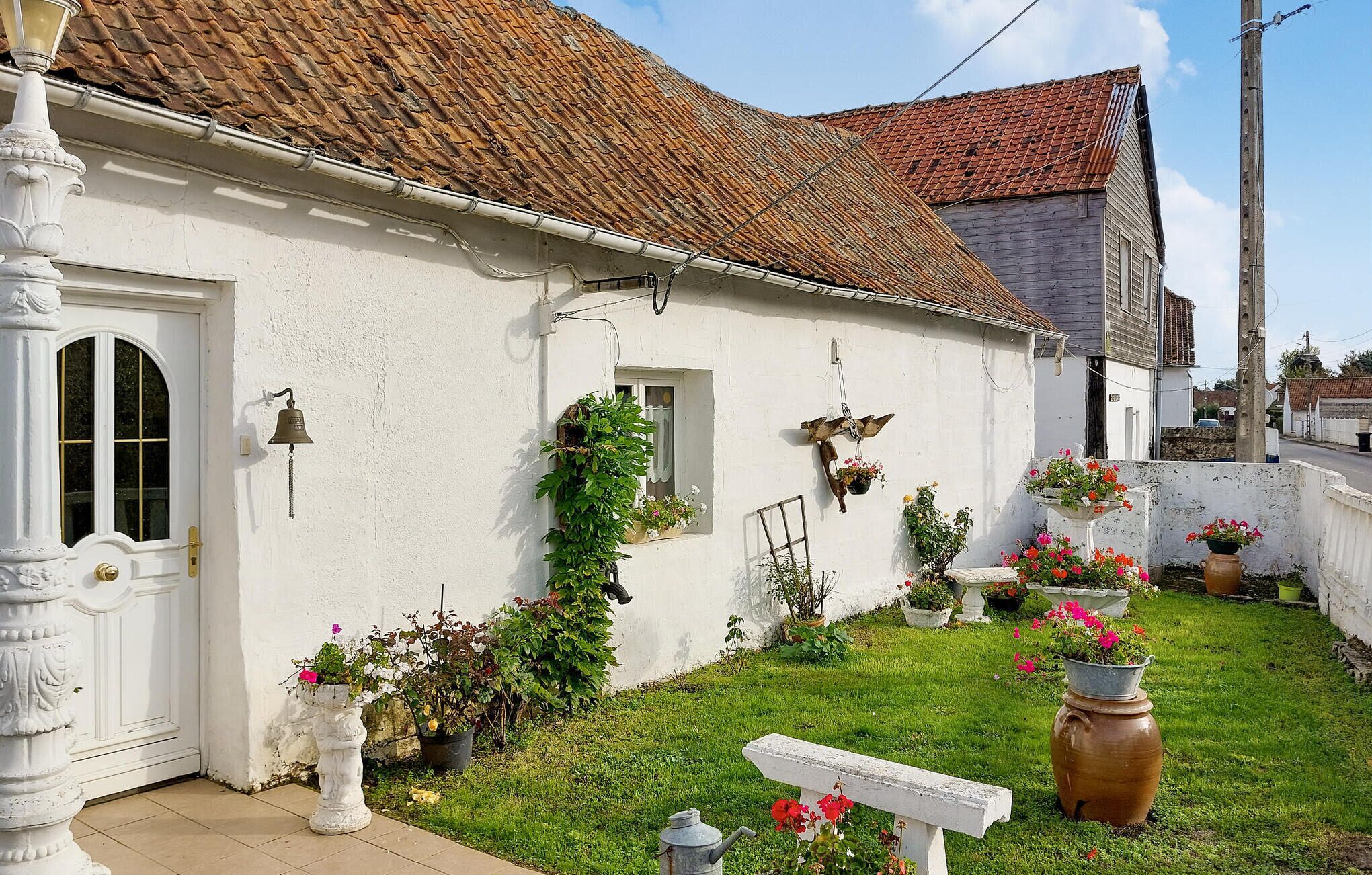 Uma casa branca com telhado de azulejos, porta da frente e uma pequena janela. Há vasos com plantas e flores no jardim.