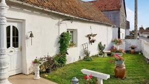 Une maison blanche avec un toit en tuiles, une porte d’entrée et une petite fenêtre. On peut voir des plantes en pot et des fleurs dans le jardin.