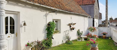 A white house with a tiled roof, a front door, and a small window. There are potted plants and flowers in the yard.