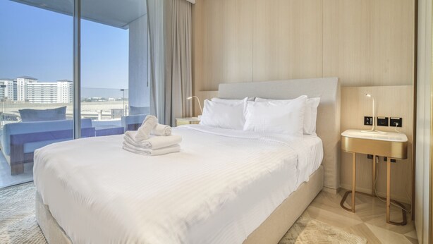 Habitació d'hotel amb un llit gran, una taula de nit i vistes de la ciutat a través de finestres de sostre a terra.