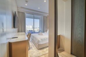 A modern hotel room with a large bed, a desk, and a view of the city through floor-to-ceiling windows.