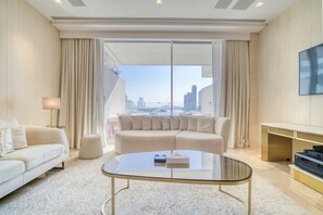 A modern hotel room with a large window offering a city view, a glass coffee table, a beige sofa, and a flat-screen TV mounted on the wall.