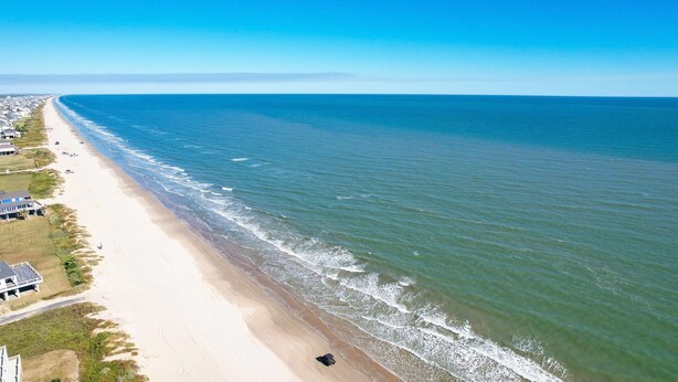 A coastal beach with a long stretch of sandy shore, a few buildings, and a clear blue sky. - S.S. Esmeralda-Newly built home-3min from beach (Bolivar Peninsula)