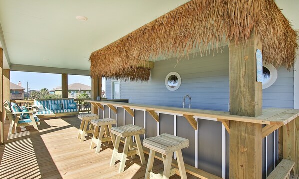 Une terrasse en bois avec un bar à toit de chaume, des tabourets en bois et une vue sur l’océan.
