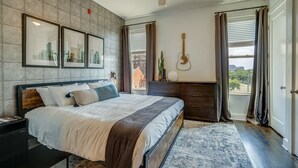 A bedroom with a bed, a nightstand, a dresser, a guitar, and a cityscape view. - Lofts at 30th-Urban Sky Suite-Walk to restaurants! (Nashville)