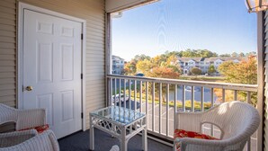 Ein Balkon mit Korbmöbeln, einem Glastisch und Blick auf eine Wohngegend mit Häusern und Bäumen.