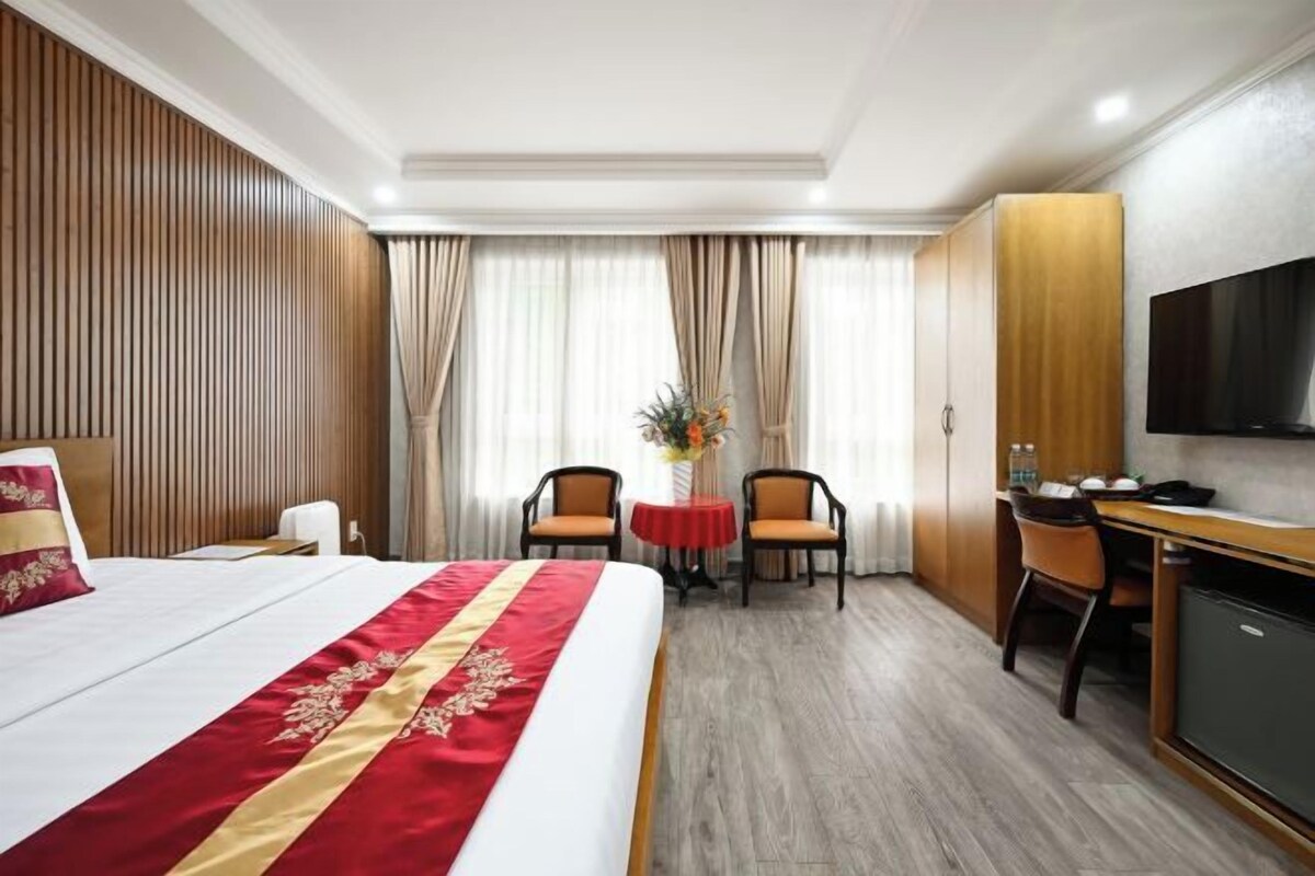 Quarto de hotel com cama, escrivaninha, duas cadeiras, televisão e pequena mesa com um vaso.