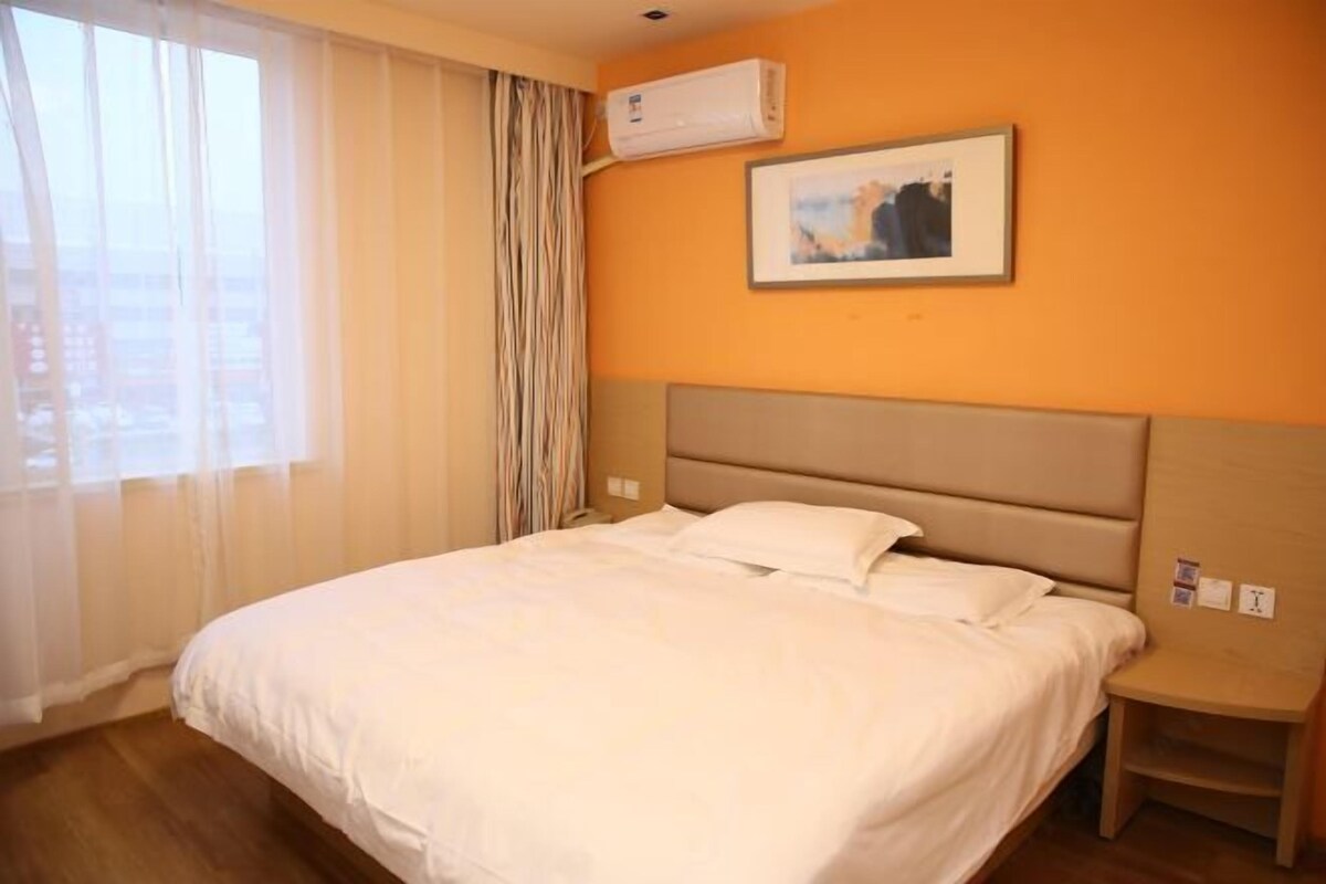 A hotel room with a bed, a bedside table, an air conditioning unit, and a window with curtains.