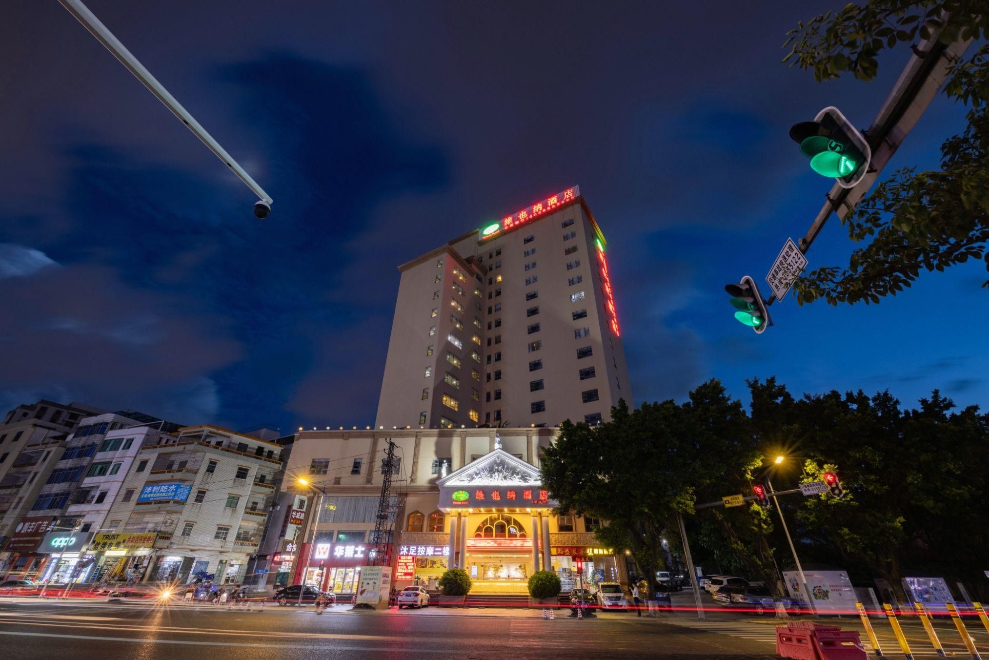 Un hôtel à plusieurs étages, doté d’une enseigne lumineuse, d’une entrée imposante surmontée d’une arche décorative, et situé sur une rue animée avec des feux de signalisation.