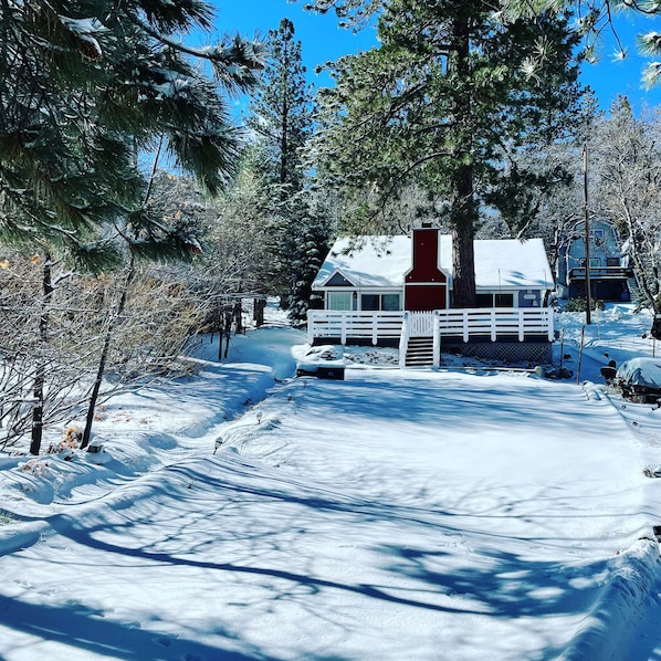 Uma cabana com chaminé vermelha, rodeada por árvores cobertas de neve e um céu claro.