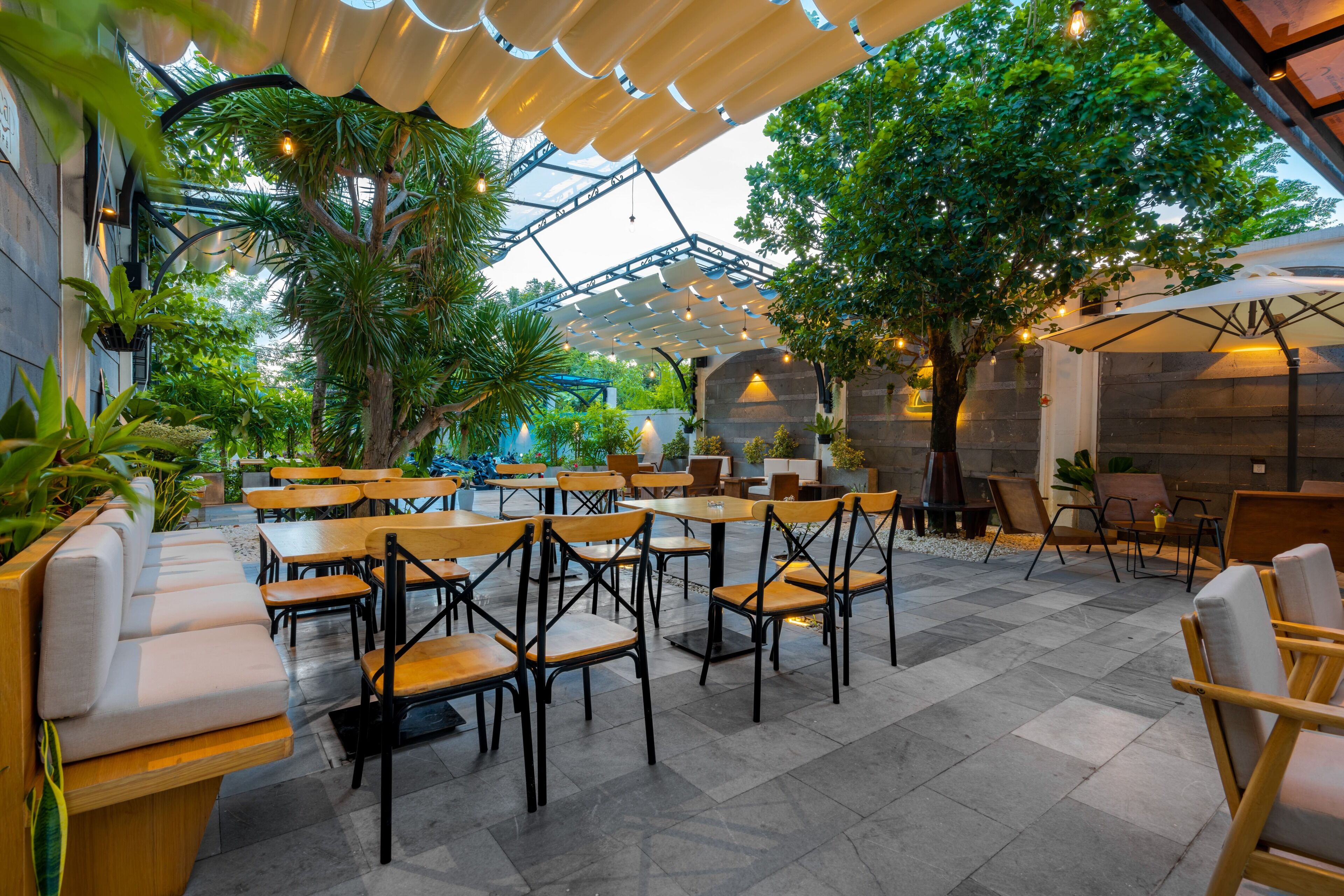 An outdoor cafe area with wooden tables and chairs, surrounded by greenery and a glass ceiling structure.