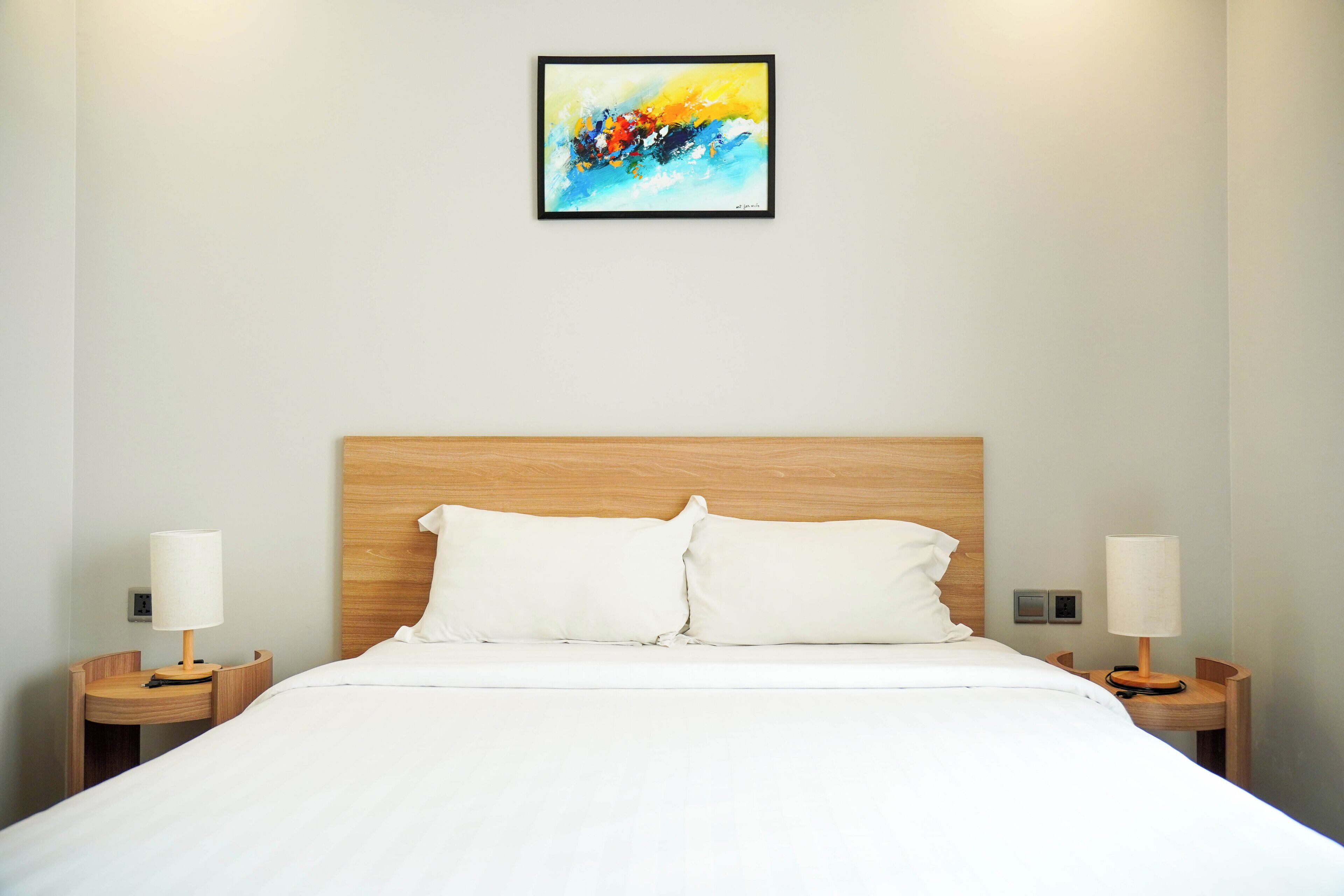 A hotel room with a wooden headboard, white bedding, two bedside tables with lamps, and a colorful abstract painting on the wall.