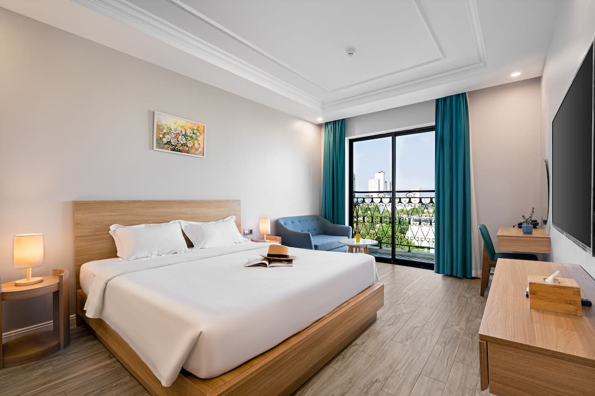 A hotel room with a large bed, a desk, a chair, a small table, a lamp, and a view of the city through a window.