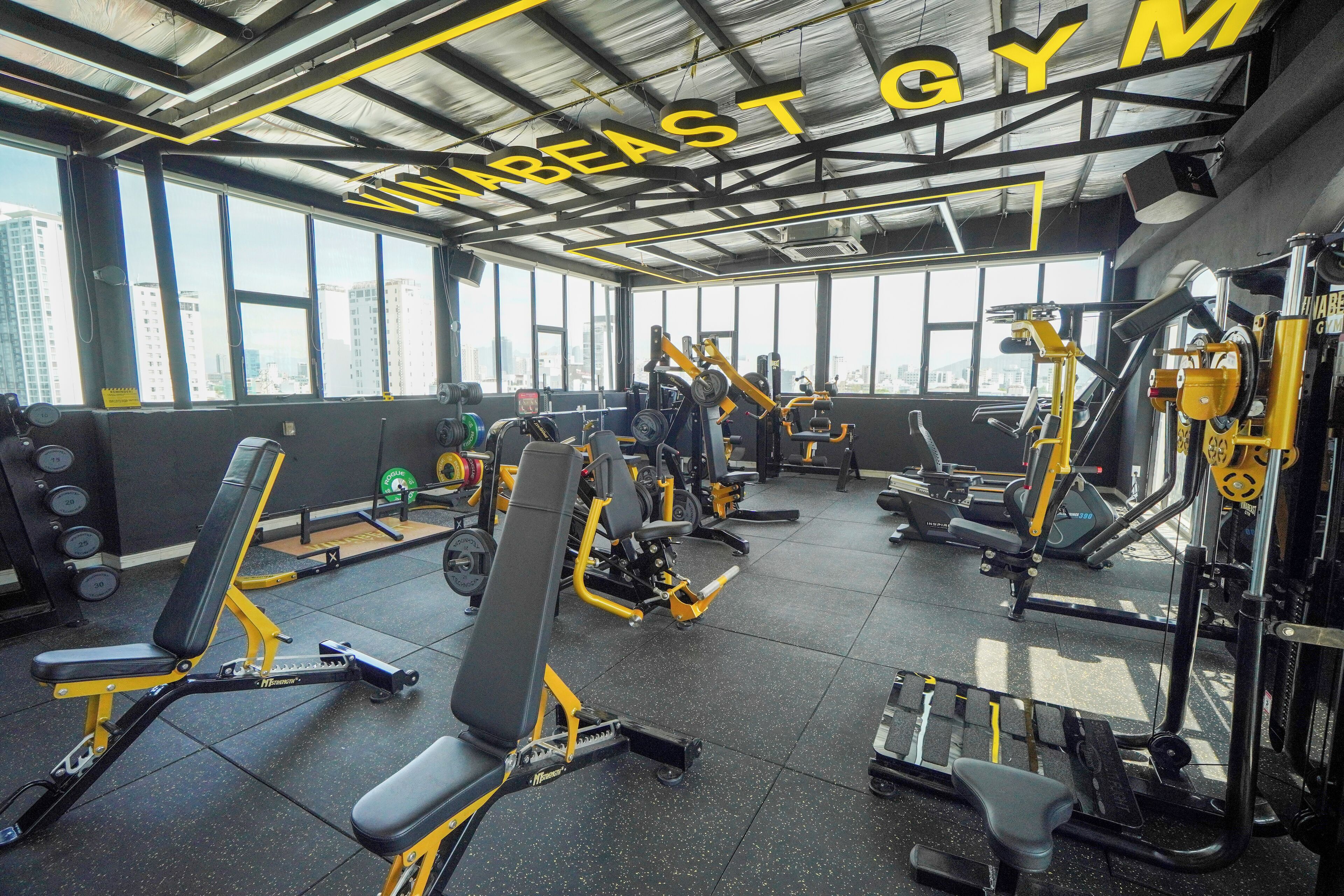 Un gimnasio con diverso equipamiento de ejercicio, ventanas amplias y la inscripción 'WREAST GYM' en el techo.