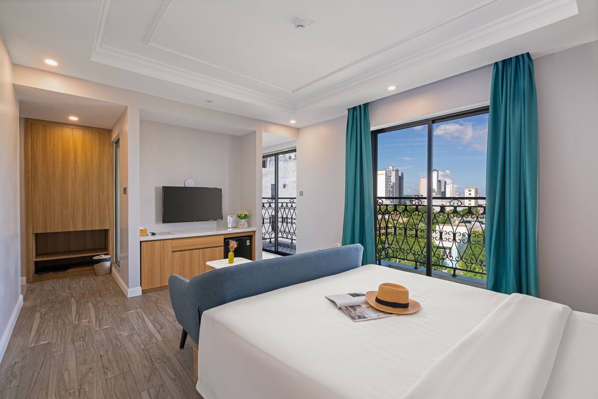 A hotel room with a bed, a desk, a TV, and a balcony with a city view.