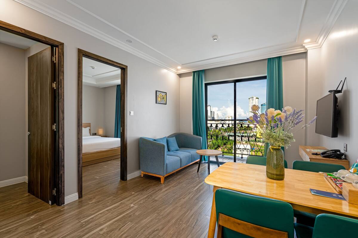 A hotel room with a bed, a sofa, a dining table, and a view of the city.