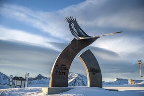 Una gran escultura de metal con inscripciones, ubicada frente a un paisaje nevado con montañas al fondo.
